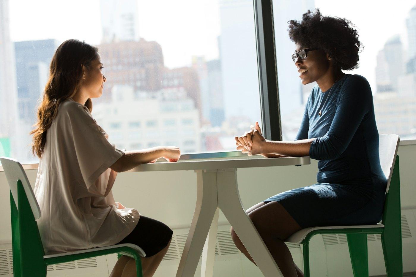 Two women talking at a table by a bright window